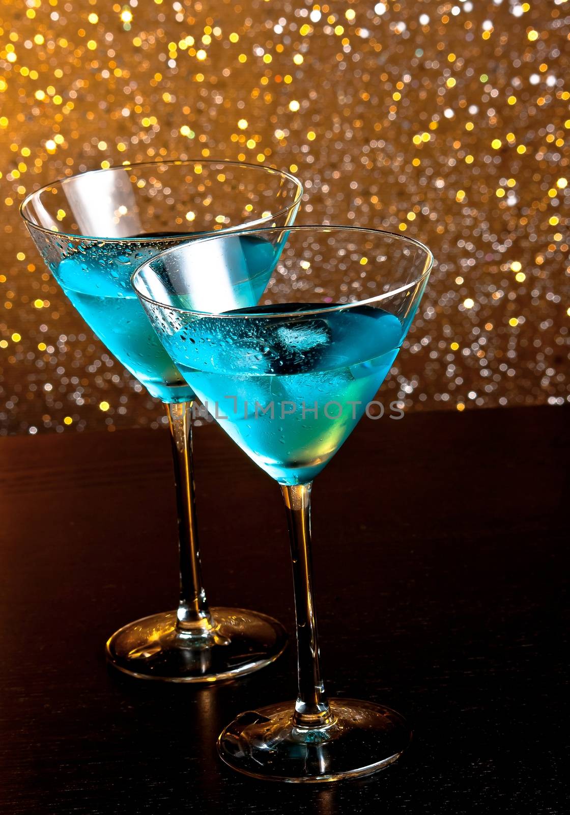 two glasses of fresh blue cocktail with ice on golden tint light bokeh background on wood table