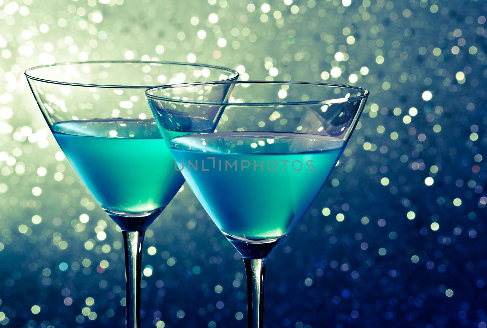 two glasses of blue cocktail on dark green tint light bokeh by donfiore