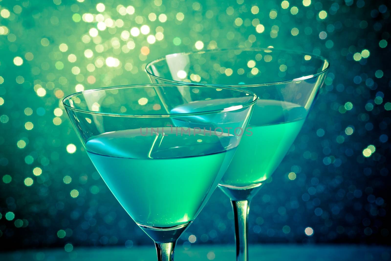 glasses of blue cocktail on dark green tint light bokeh by donfiore