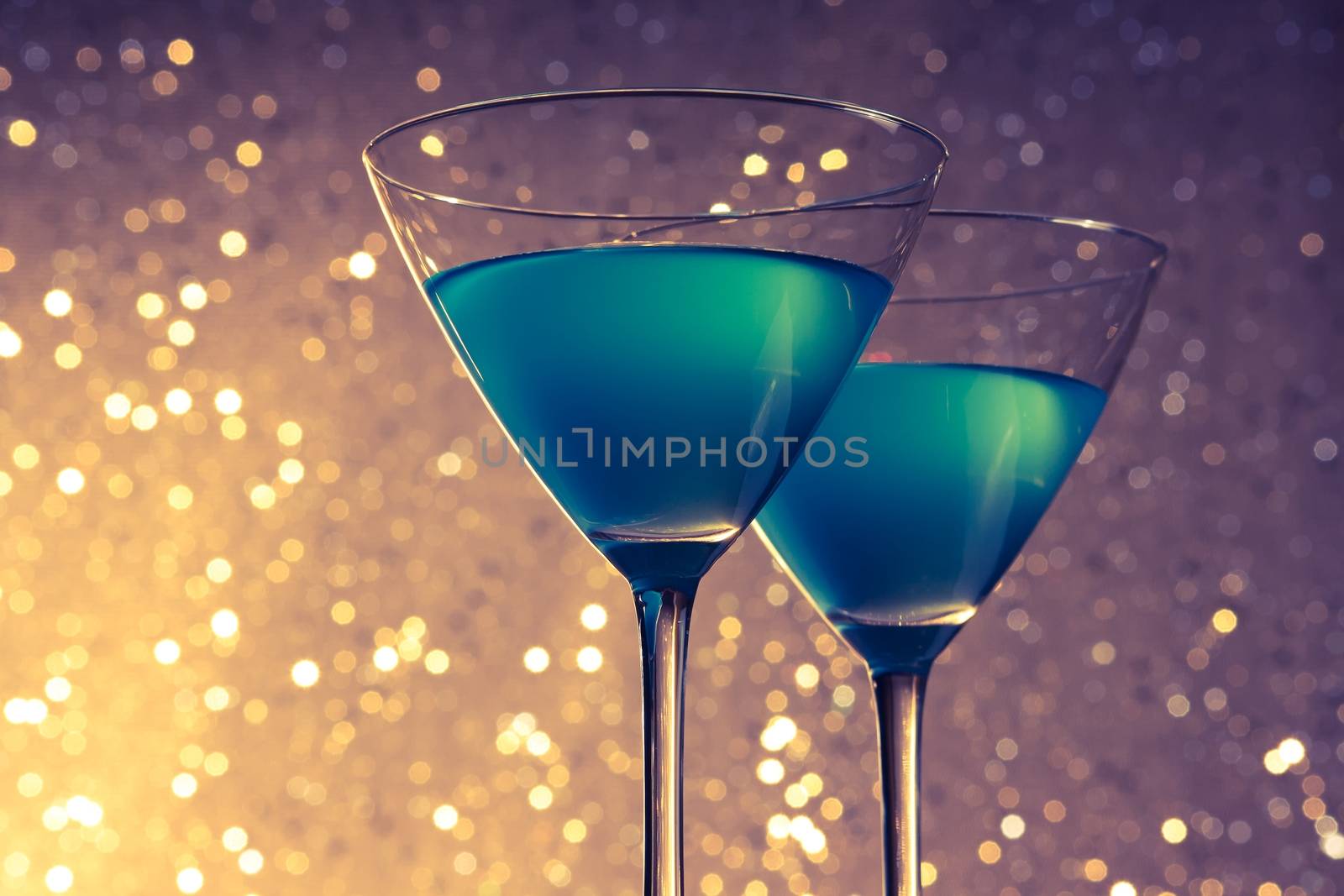 glasses of blue cocktail on golden and violet tint light bokeh background on table