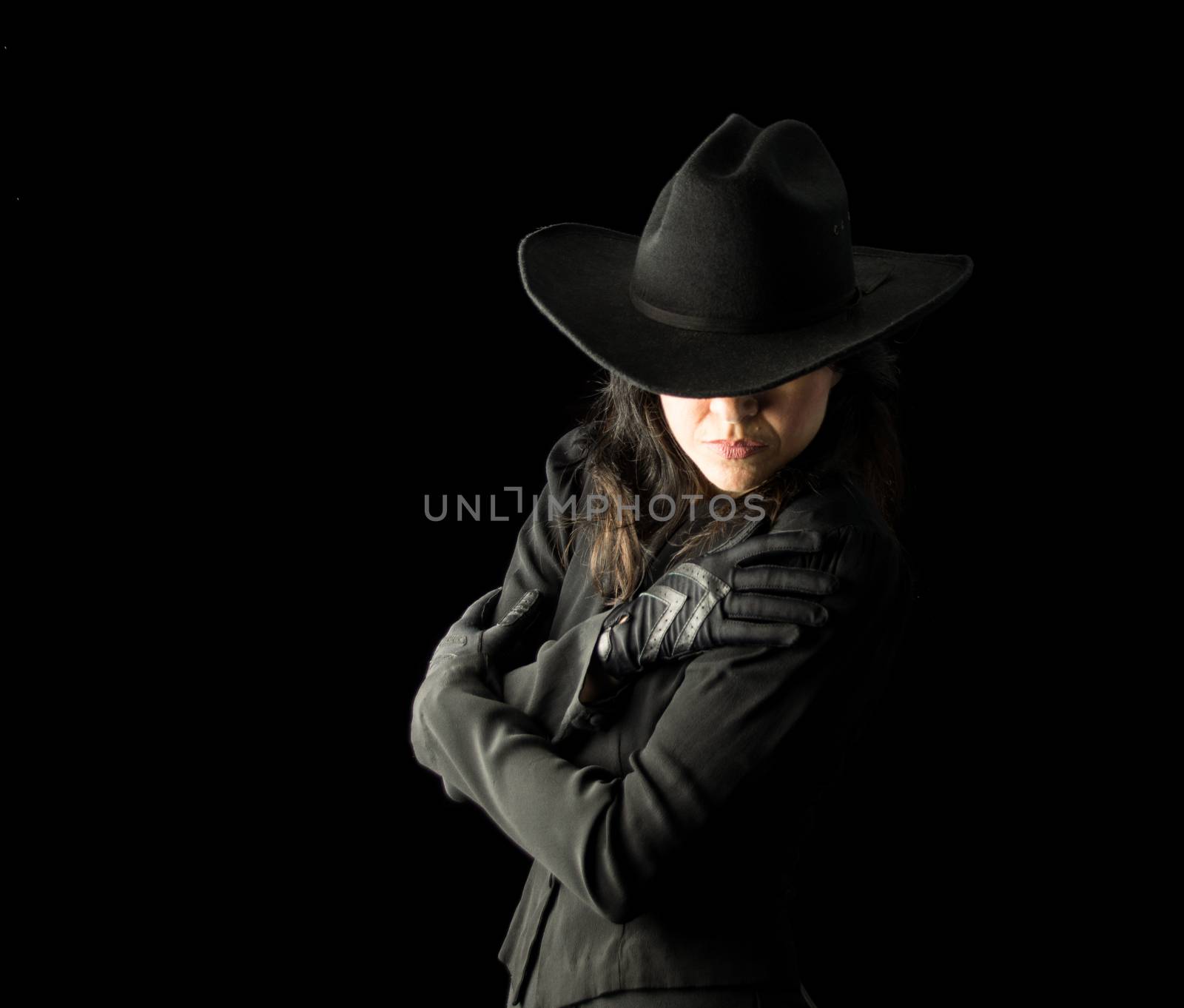 Brunette woman in black dress and black gloves, wearing a black cowboy hat, holding her arms across her body and looking downward, with her face partially hidden by the hat.