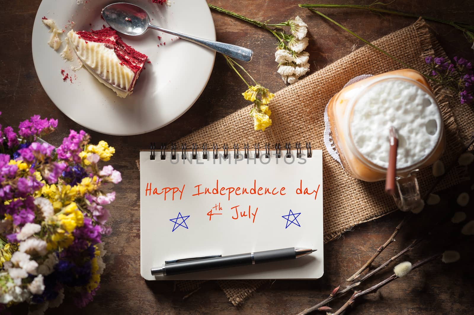 "Happy independence day 4th july" is written on Notepad with pen, a glass of Thai tea and bits of red velvet cake on rustic wood background with low key scene