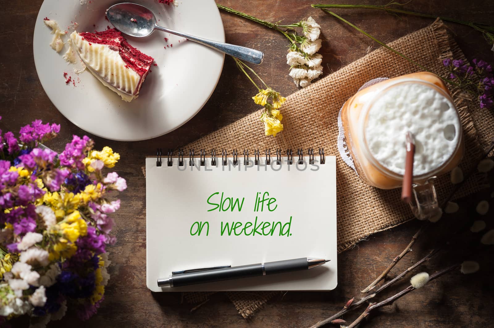 "Slow life on weekend" is written on Notepad with pen, a glass of Thai tea and bits of red velvet cake on rustic wood background with low key scene