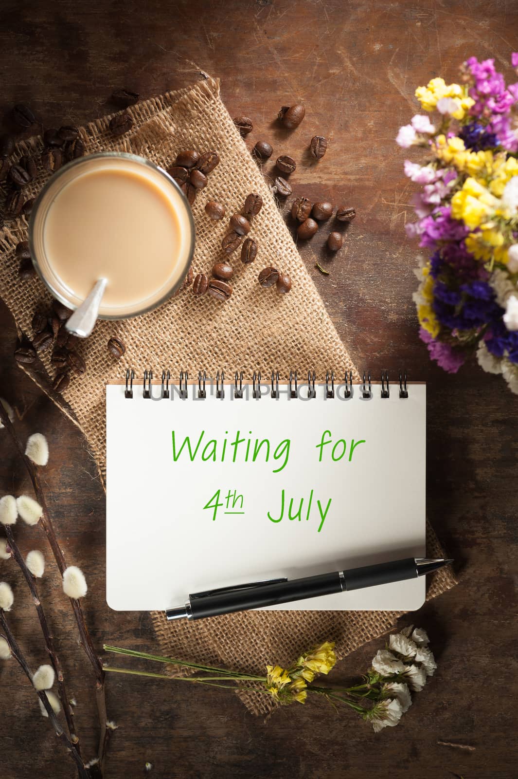 "waiting for 4th july" is written on Notepad with pen, a glass of Thai tea and bits of red velvet cake on rustic wood background with low key scene
