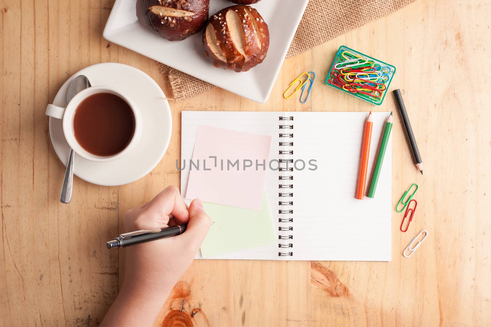 Notebook and sticky paper, paper clip, color pencil, and pen on wood table in vintage style with female left hand wrting on notepad