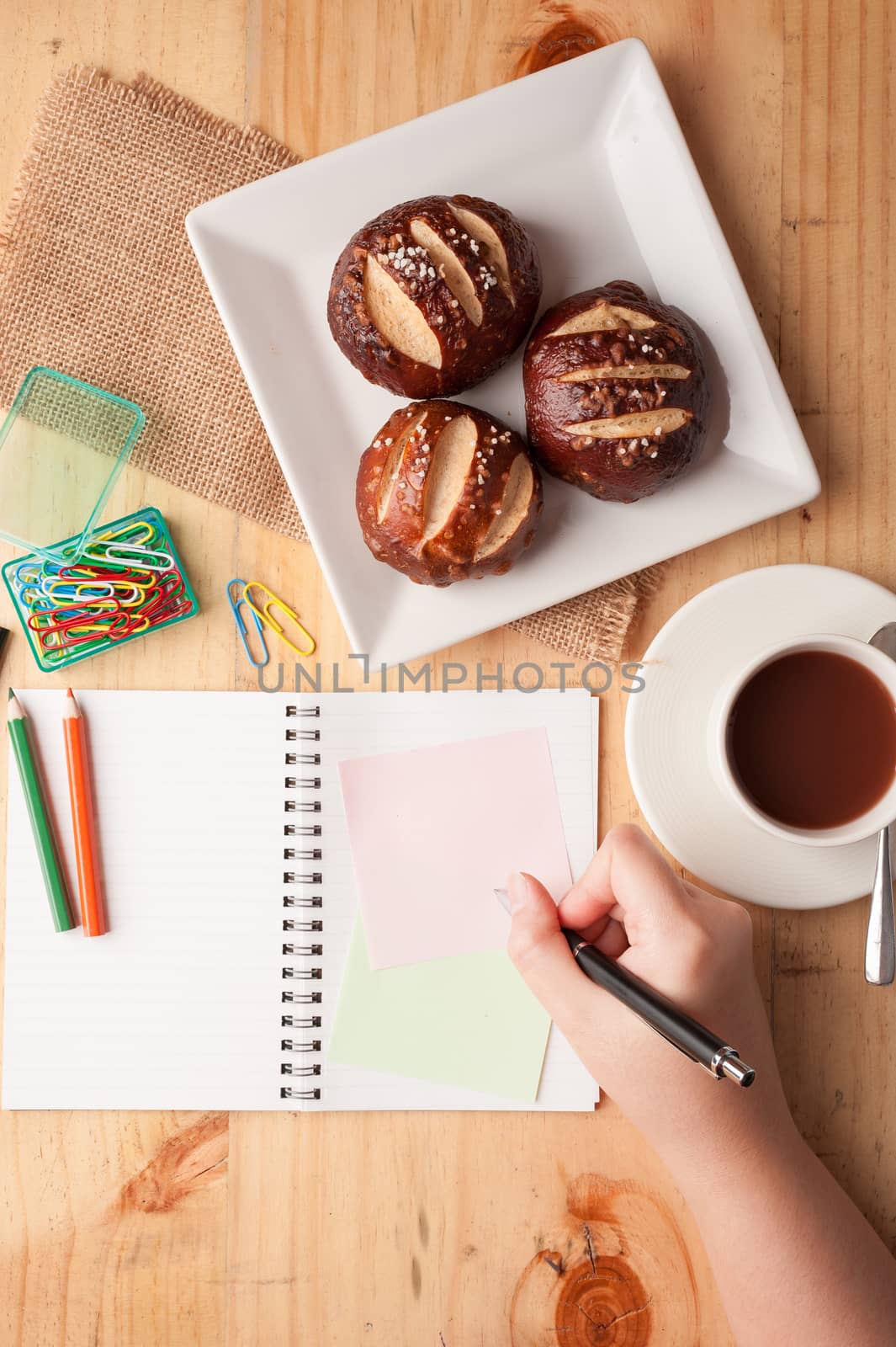 Notebook and sticky paper, paper clip, color pencil, and pen on wood table in vintage style with female right hand wrting on notepad