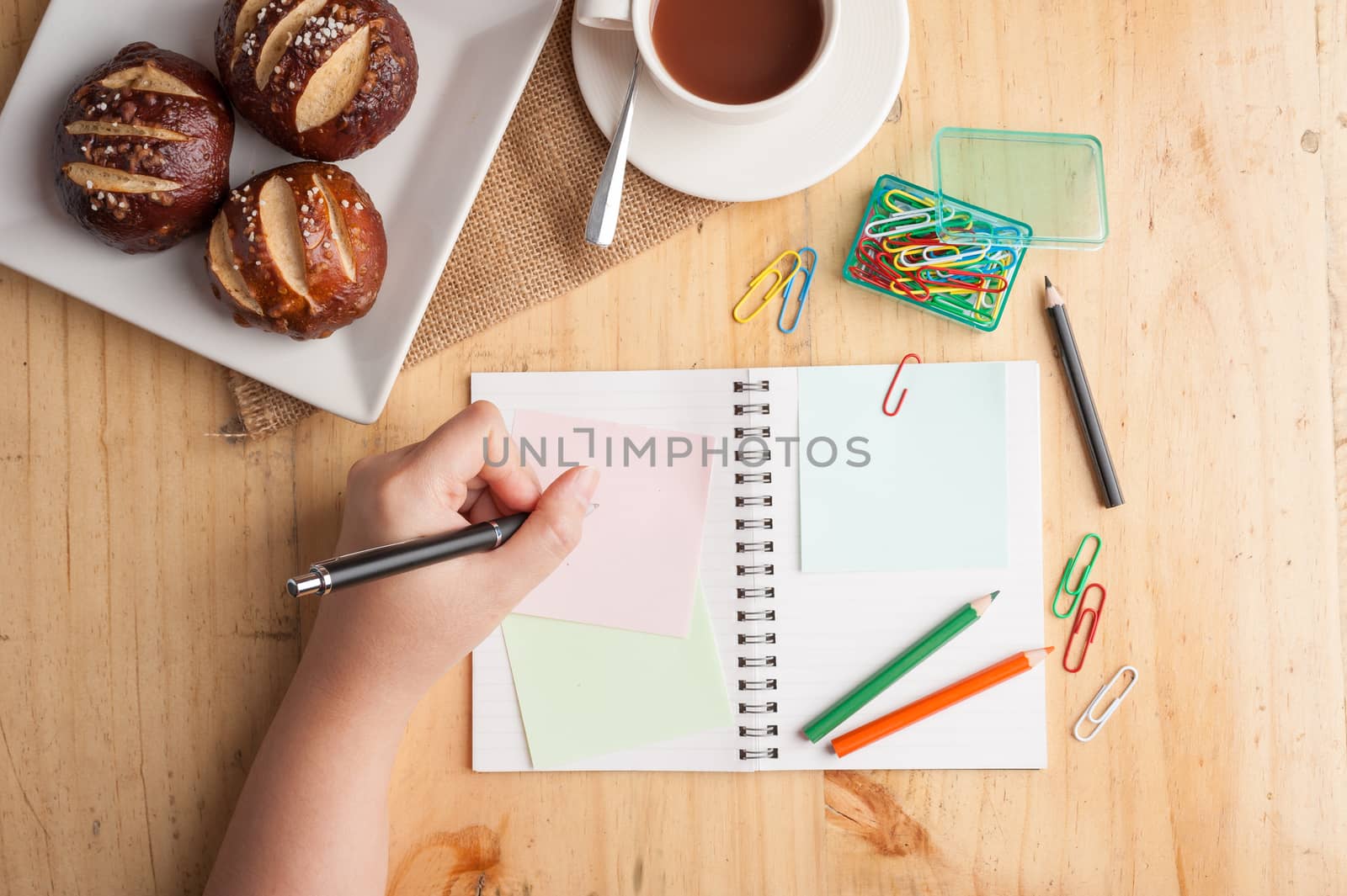 Notebook and sticky paper, paper clip, color pencil, and pen on wood table in vintage style with female hand wrting on notepad