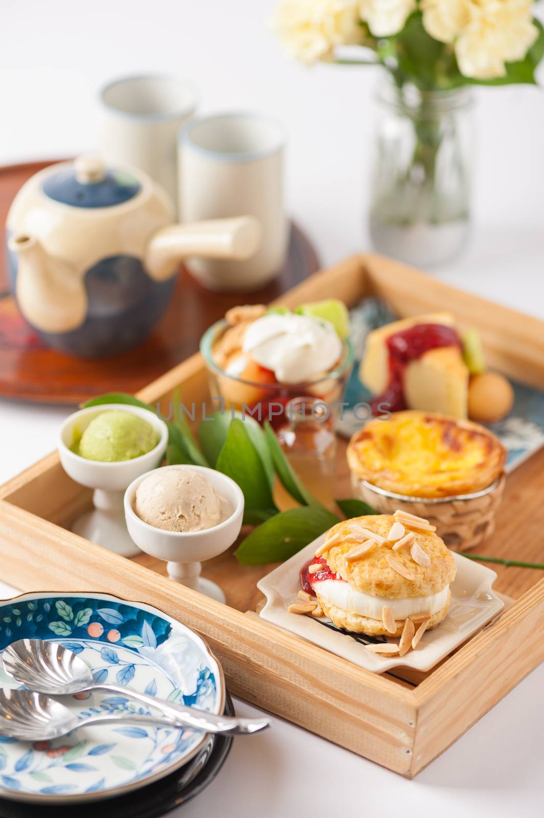 Dessert set for tea time with cake, ice cream, egg tart, and scone in wood tray on white table with tea set and diashes