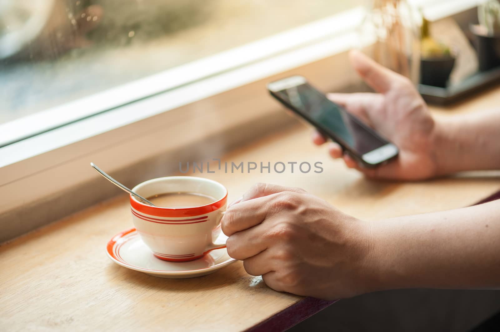 Male hand holding a cup of coffee on wood bar with another hand using smartphone in blurry background with morning scene