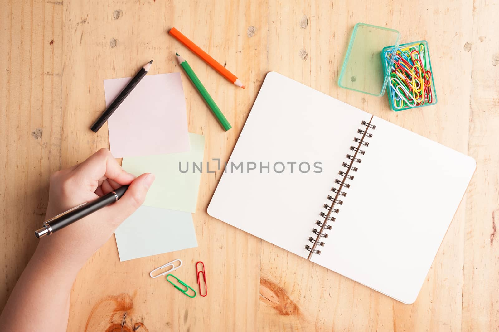 Notebook and sticky paper, paper clip, color pencil, and pen with female left hand on wood table