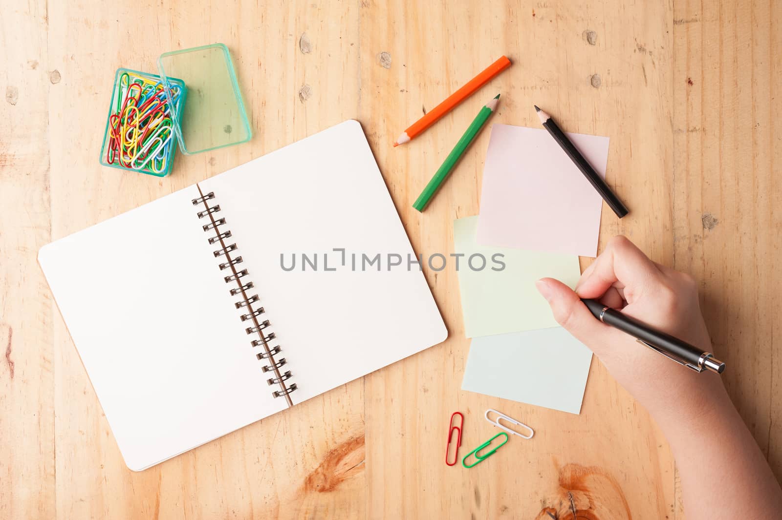 Notebook and sticky paper, paper clip, color pencil, and pen with female right hand on wood table