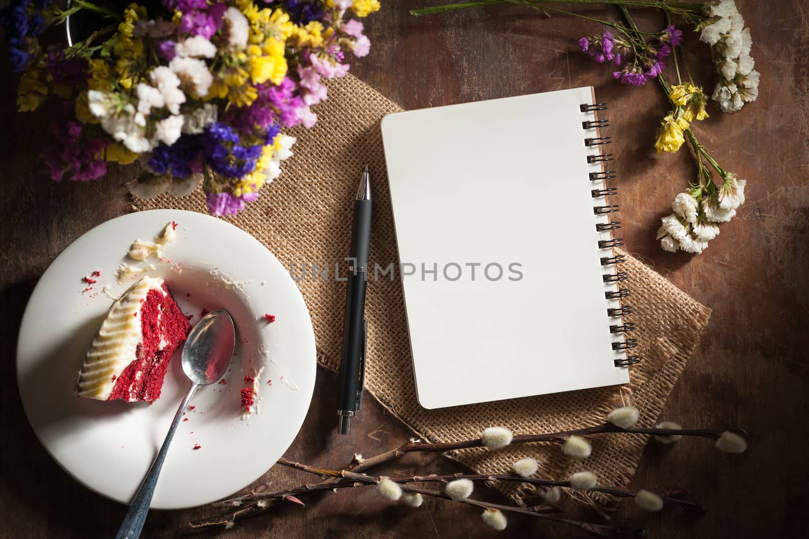 Small notepad with pen, pencil and bits of red velvet cake on rustic wood background with low key scene.