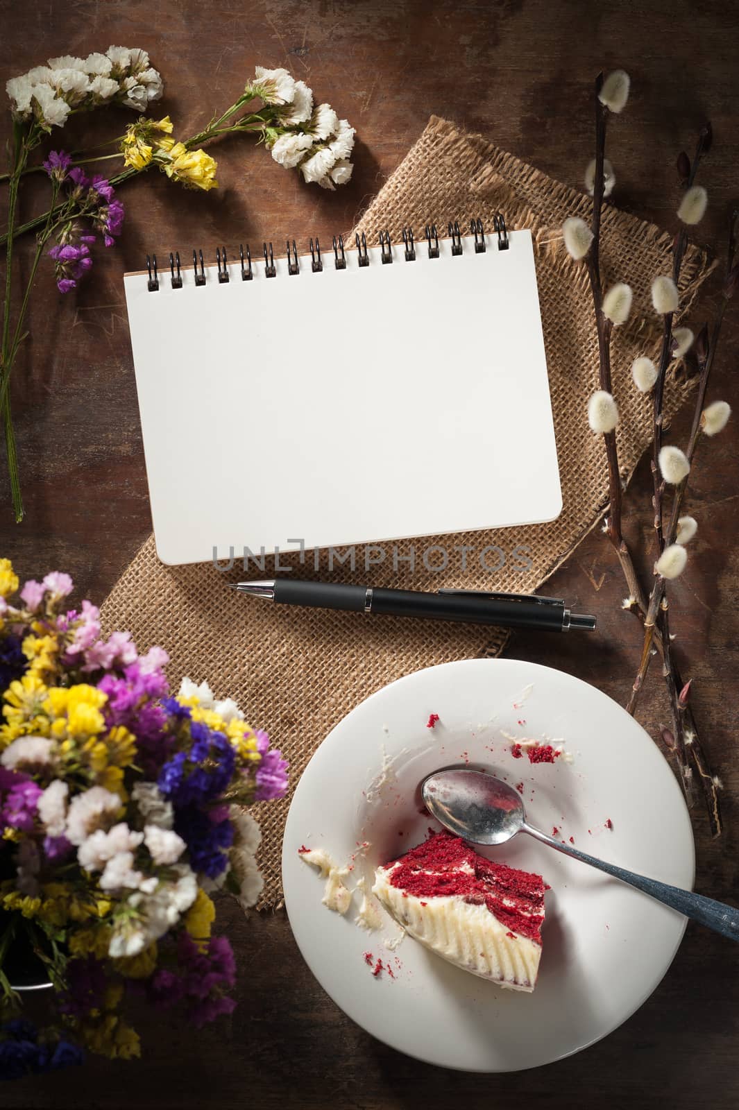 Small notepad with pen, pencil and bits of red velvet cake on rustic wood background with low key scene.