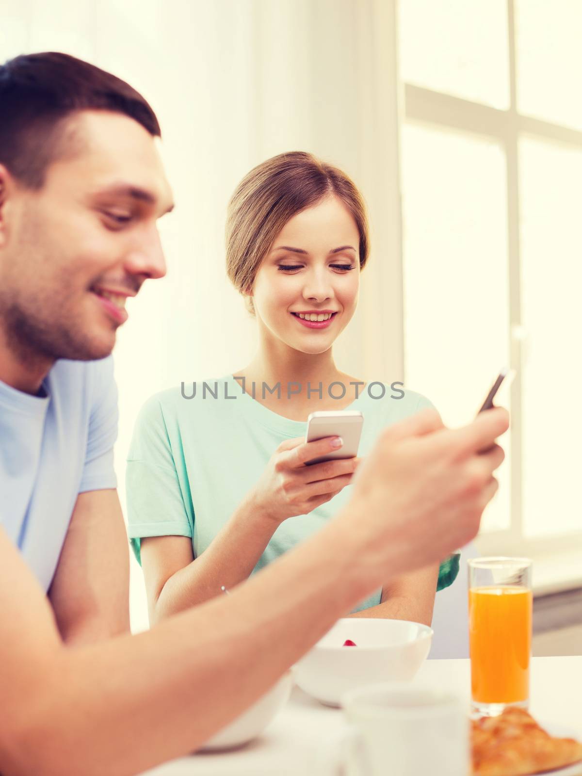 food, home, couple and technology concept - smiling couple with smartphones reading news and having breakfast at home