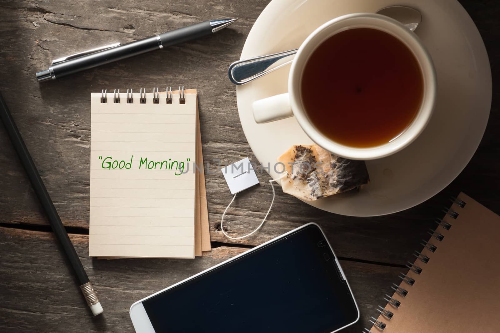 "Good morning" is written on small notepad with a cup of tea, pen, pencil and cellphne on rustic wood background with low key scene