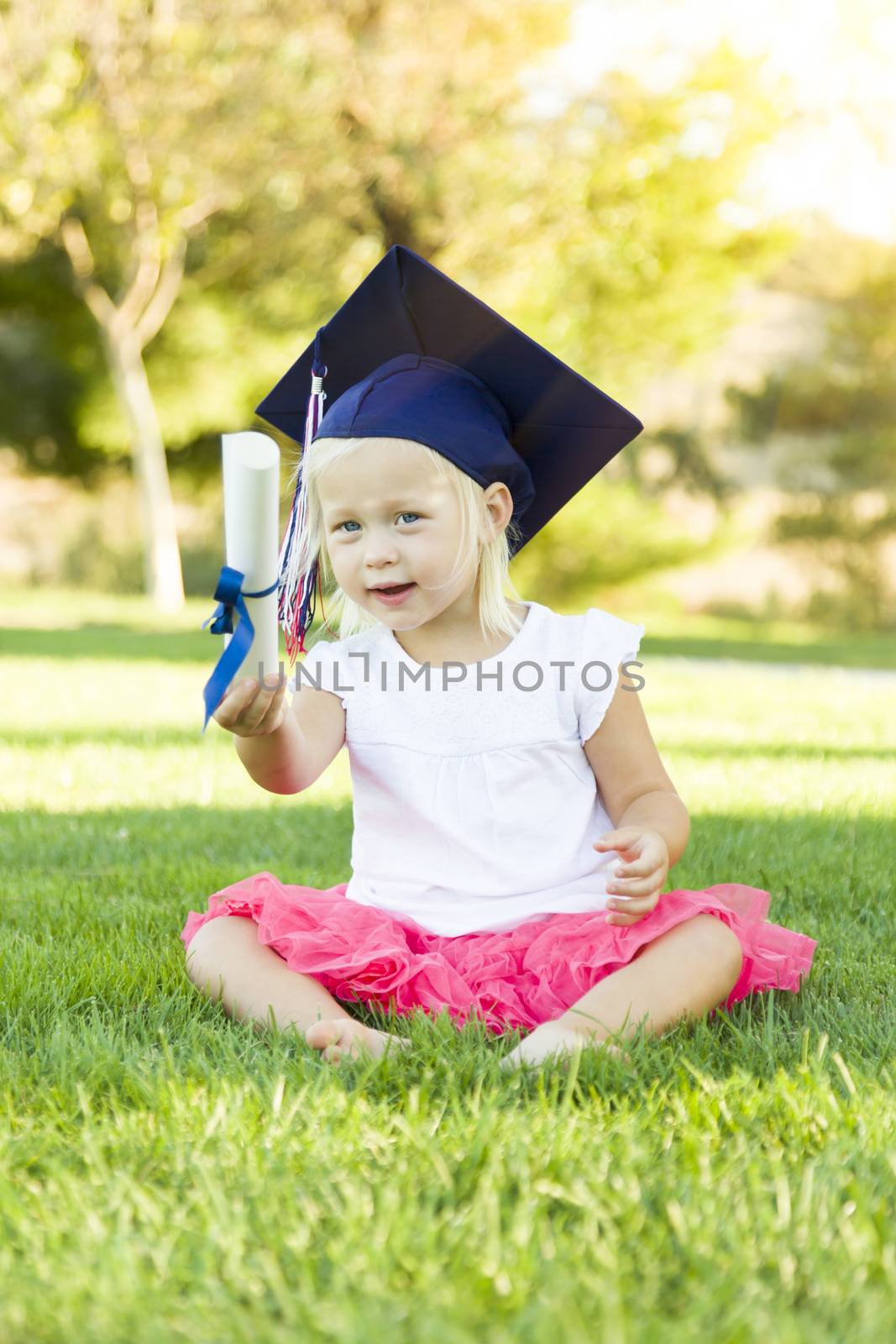 Cute Little Girl In Grass Wearing Graduation Cap Holding Diploma With Ribbon.