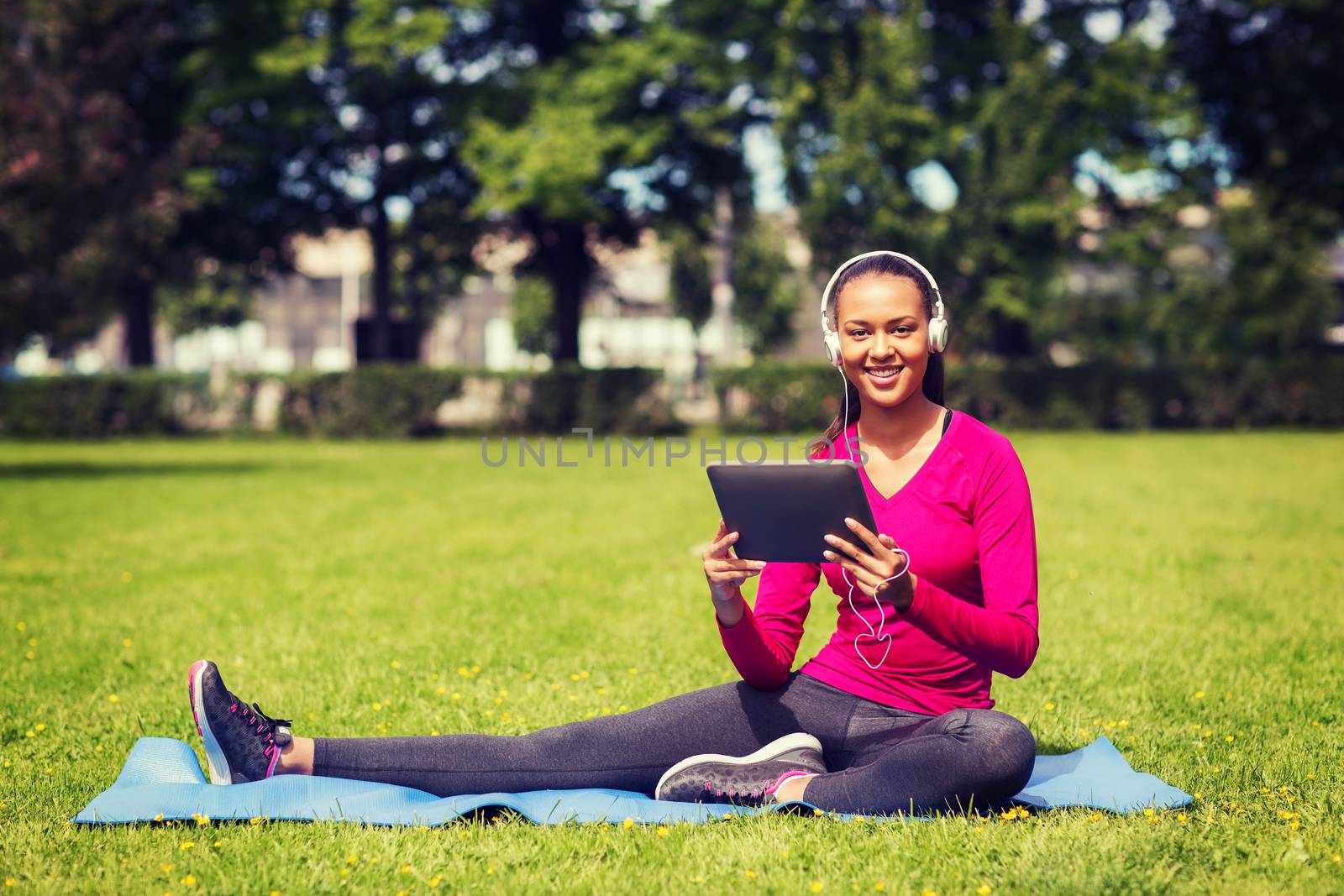 fitness, park, technology and sport concept - smiling african american woman with tablet pc computer and headphones on mat outdoors