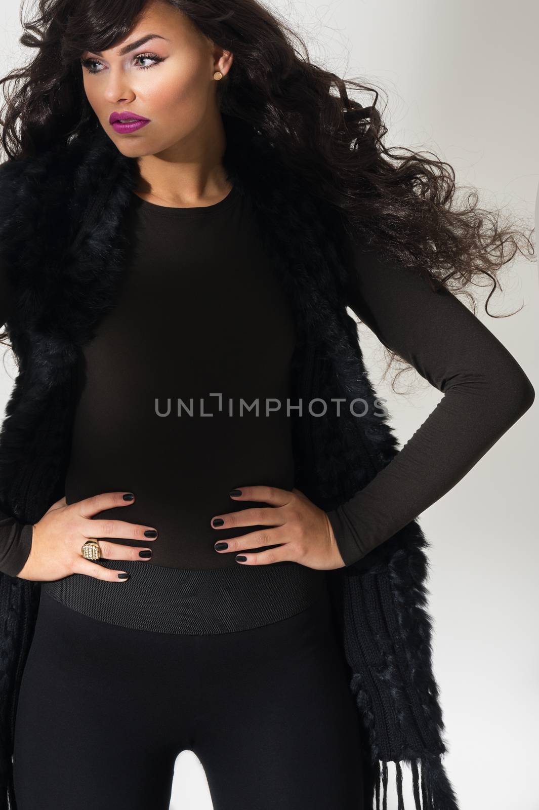 Confident attractive young fashion model with flowing long curly auburn hair standing with her hands on her hips in an elegant black ensemble, close up view over grey
