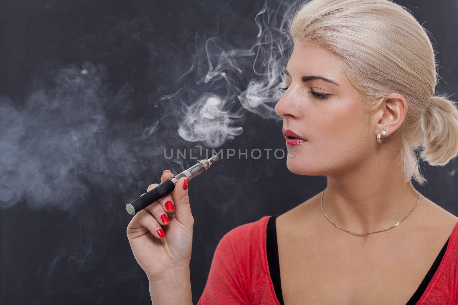 Stylish blond woman smoking an e-cigarette exhaling a cloud of smoke with her eyes closed in enjoyment, profile view on a dark background