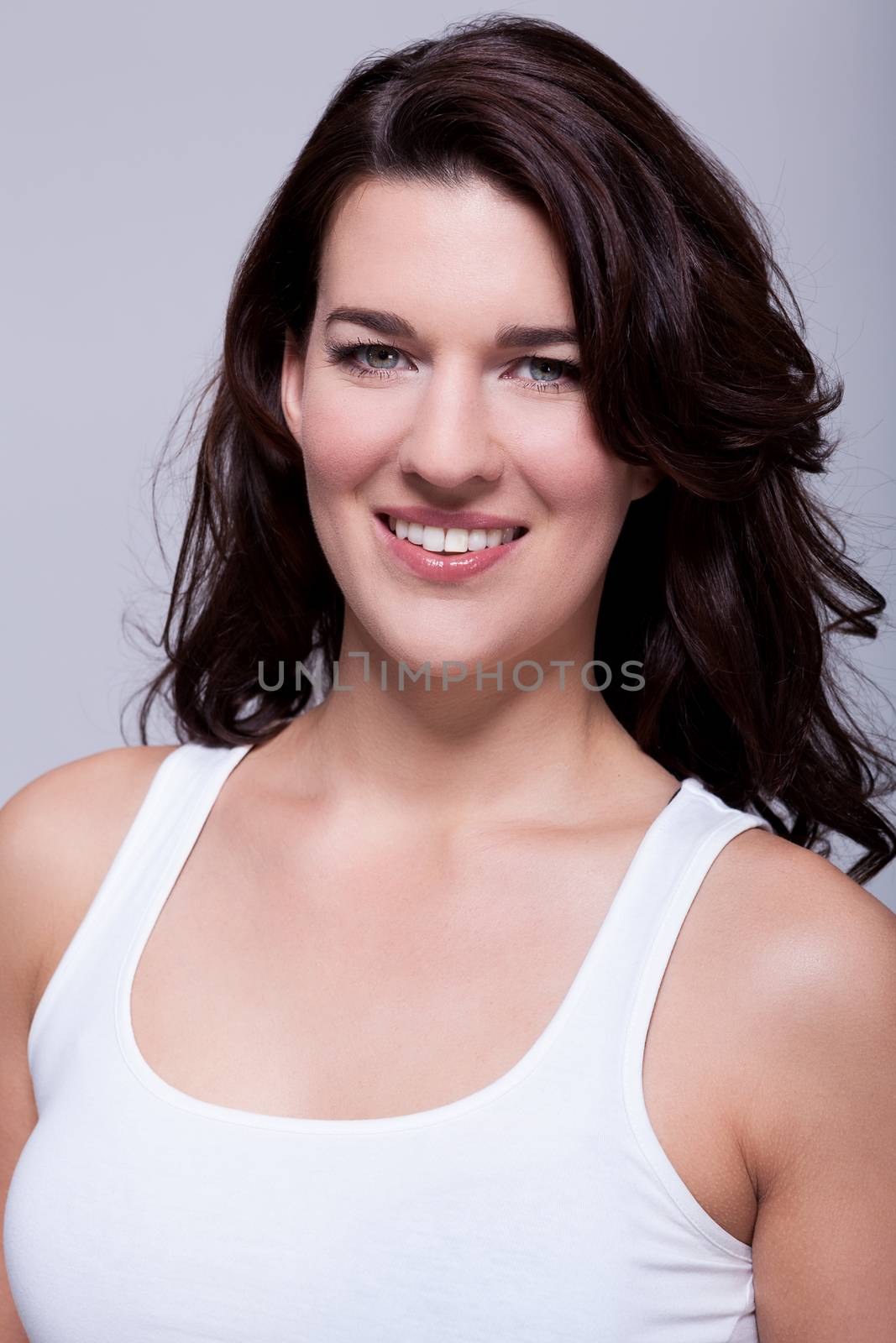 Smiling attractive woman with a lovely warm friendly smile and shoulder length curly brown hair looking at the camera over a grey background