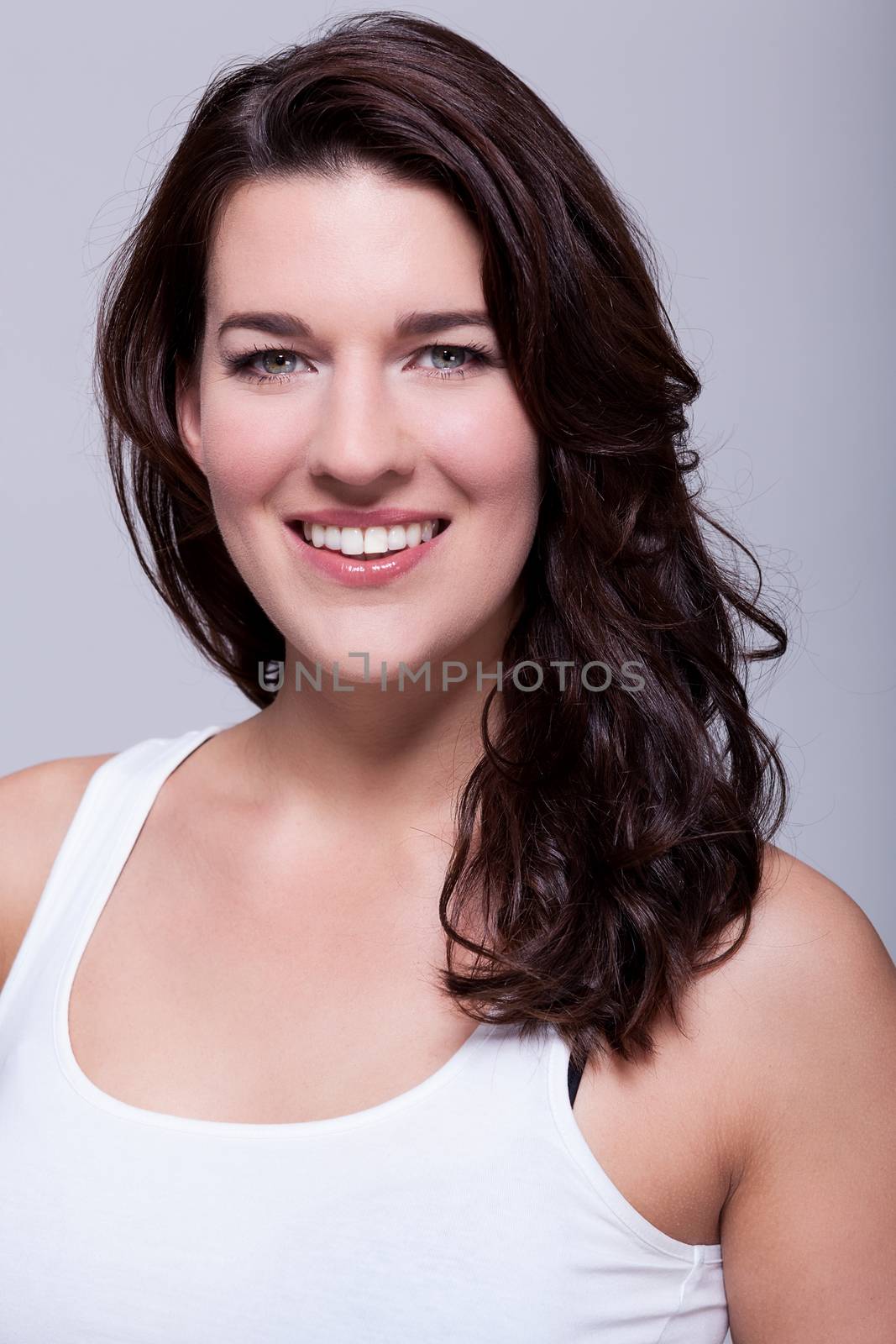 Smiling attractive woman with a lovely warm friendly smile and shoulder length curly brown hair looking at the camera over a grey background