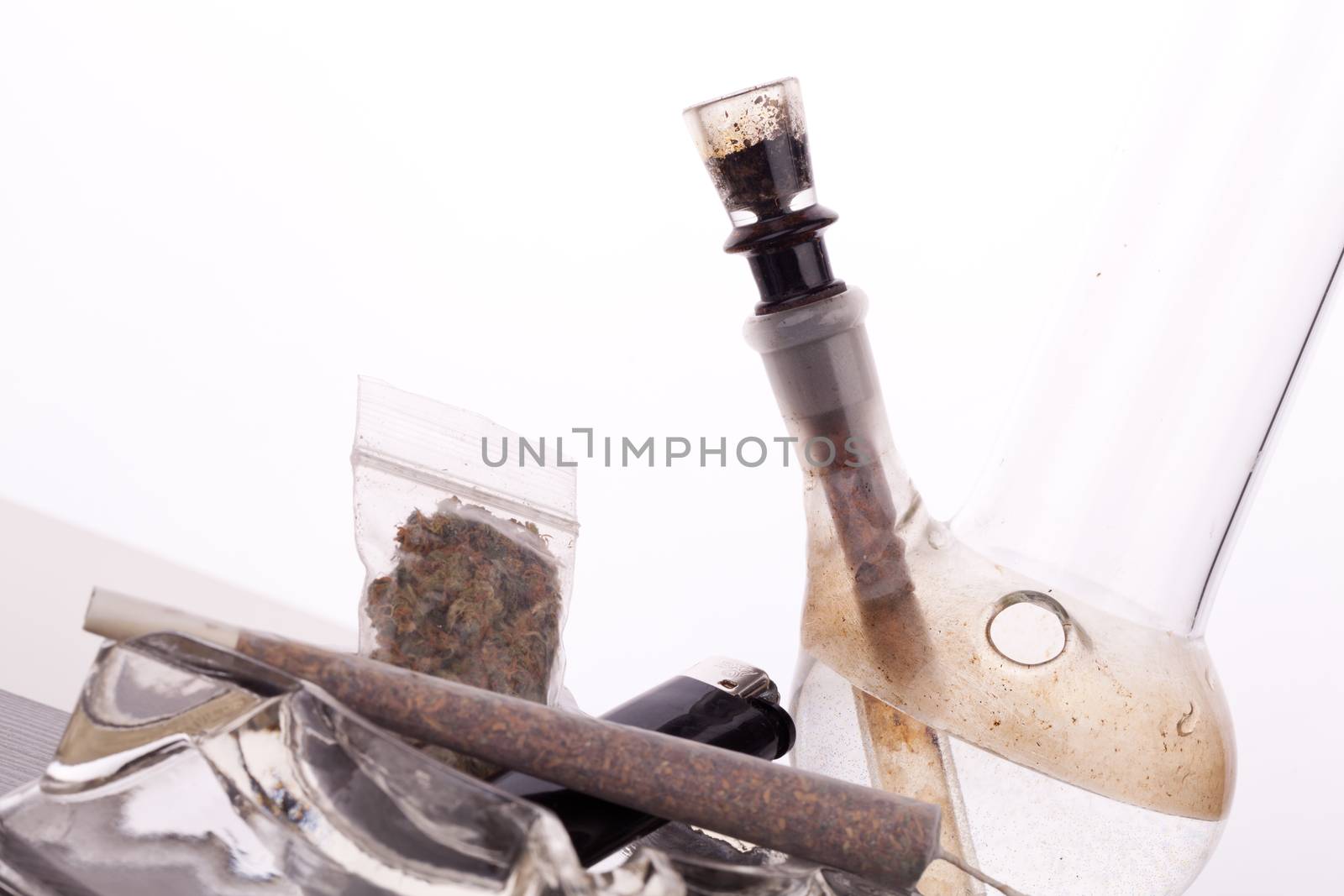 Close up of marijuana joint made with translucent rolling papers, plastic baggy of dried marijuana, black lighter and pipe on white background