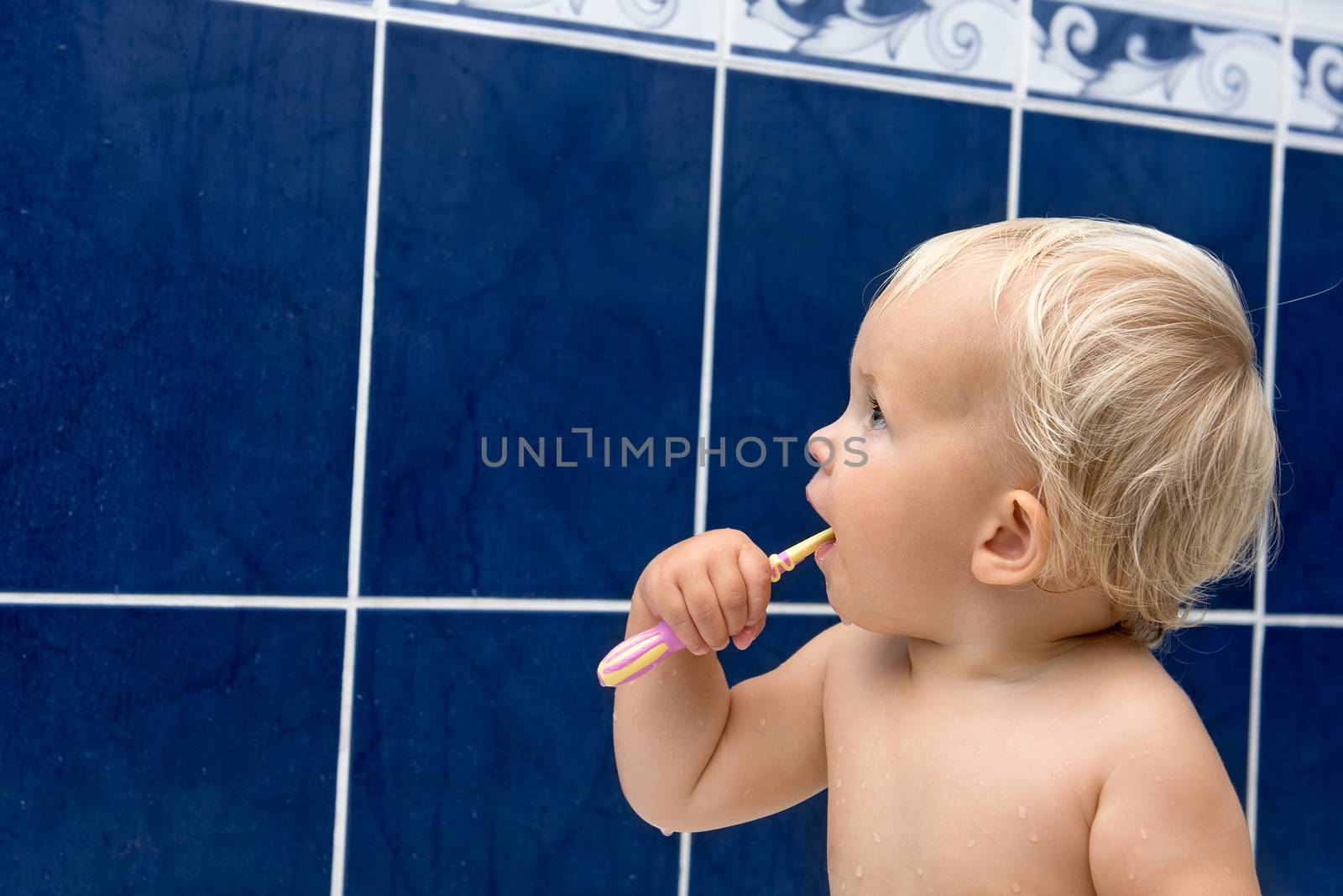Little girl is brushing teeth in bathroom. Blue tiles behind. Looking sideways
