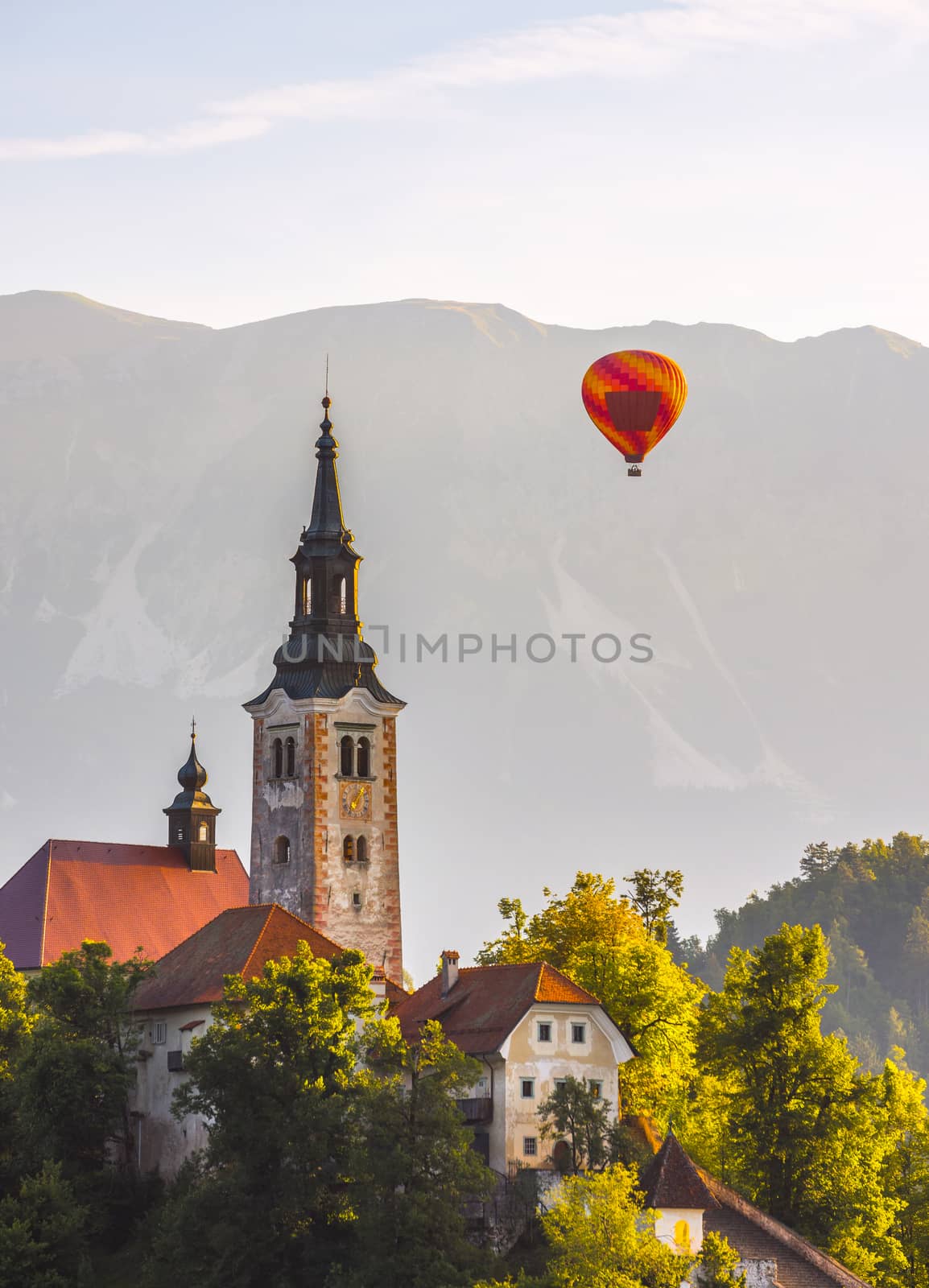 Detail of Famous Catholic Church in Bled Lake, Slovenia with Hot Air Balloon Flying with Mountains in Background at Sunrise