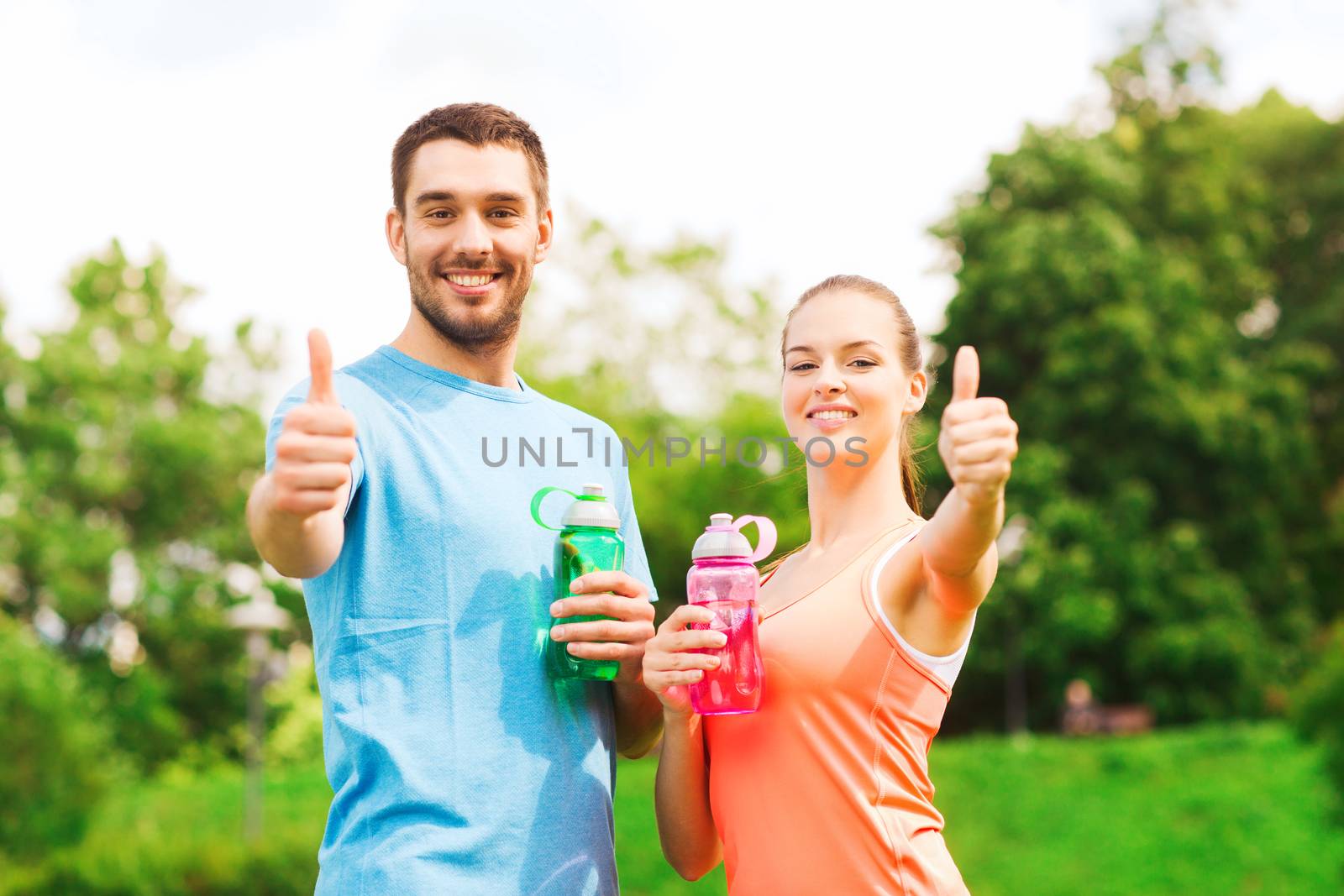 fitness, sport, friendship and lifestyle concept - smiling couple with bottles of water showing thumbs up outdoors