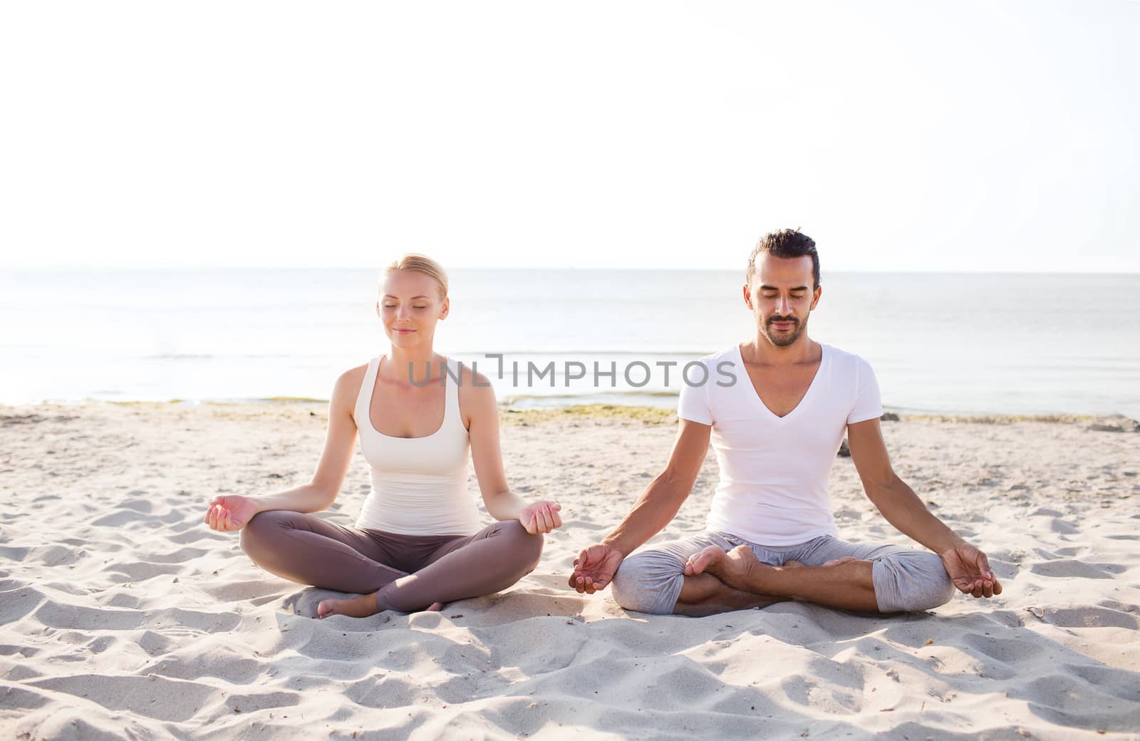 fitness, sport, friendship and lifestyle concept - smiling couple making yoga exercises sitting on sand outdoors