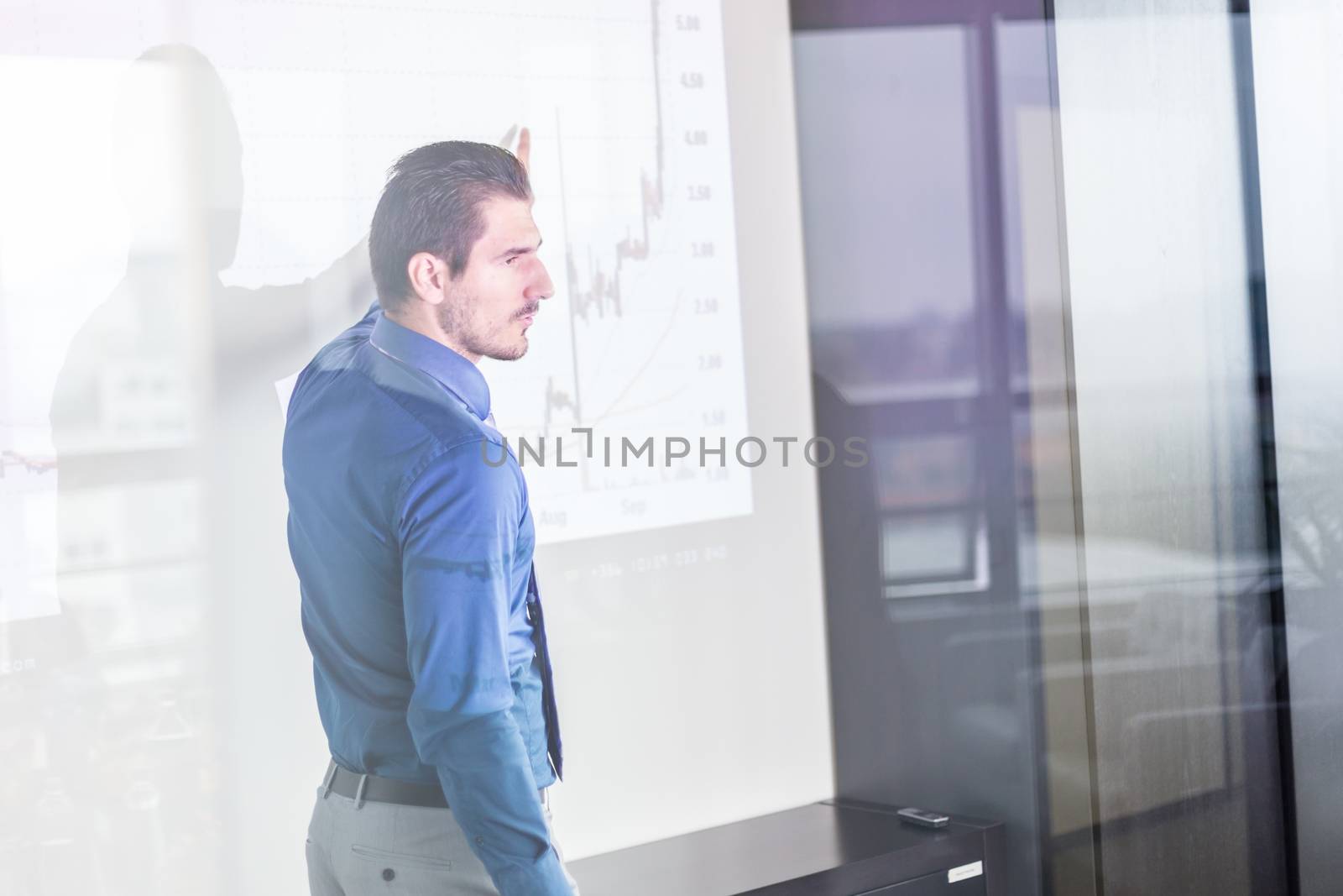 Business man making a presentation in front of whiteboard. Business executive delivering a presentation to his colleagues during meeting or in-house business training. View through glass.