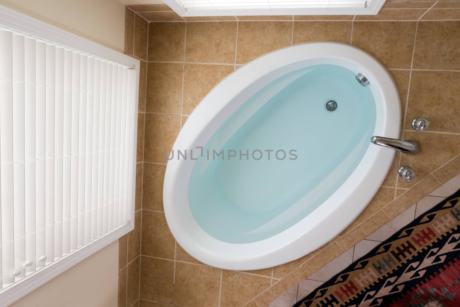 Modern oval domestic bathtub full of clean water in a brown tile surround below a tall window with blinds viewed from above