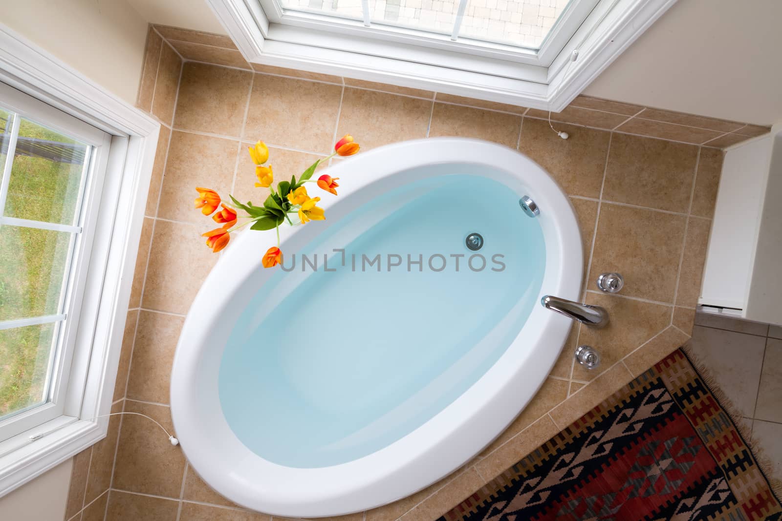 Corner oval bathtub full of clean water sunken into a brown tile surround and flanked by two tall view windows, overhead view