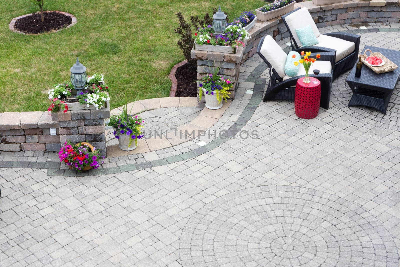 Decorative brick paving on an outdoor patio with a circular pattern and curved steps flanked by spring flowers leading to a green lawn, comfortable seating in the background