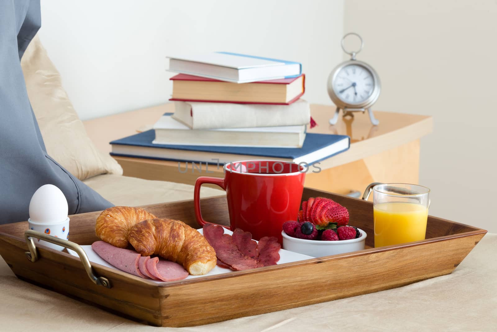 Close Up of Breakfast in Bed, Wooden Tray with Assortment of Appetizing Food Resting on Bed Next to Bedside Table Stacked with Books and Alarm Clock