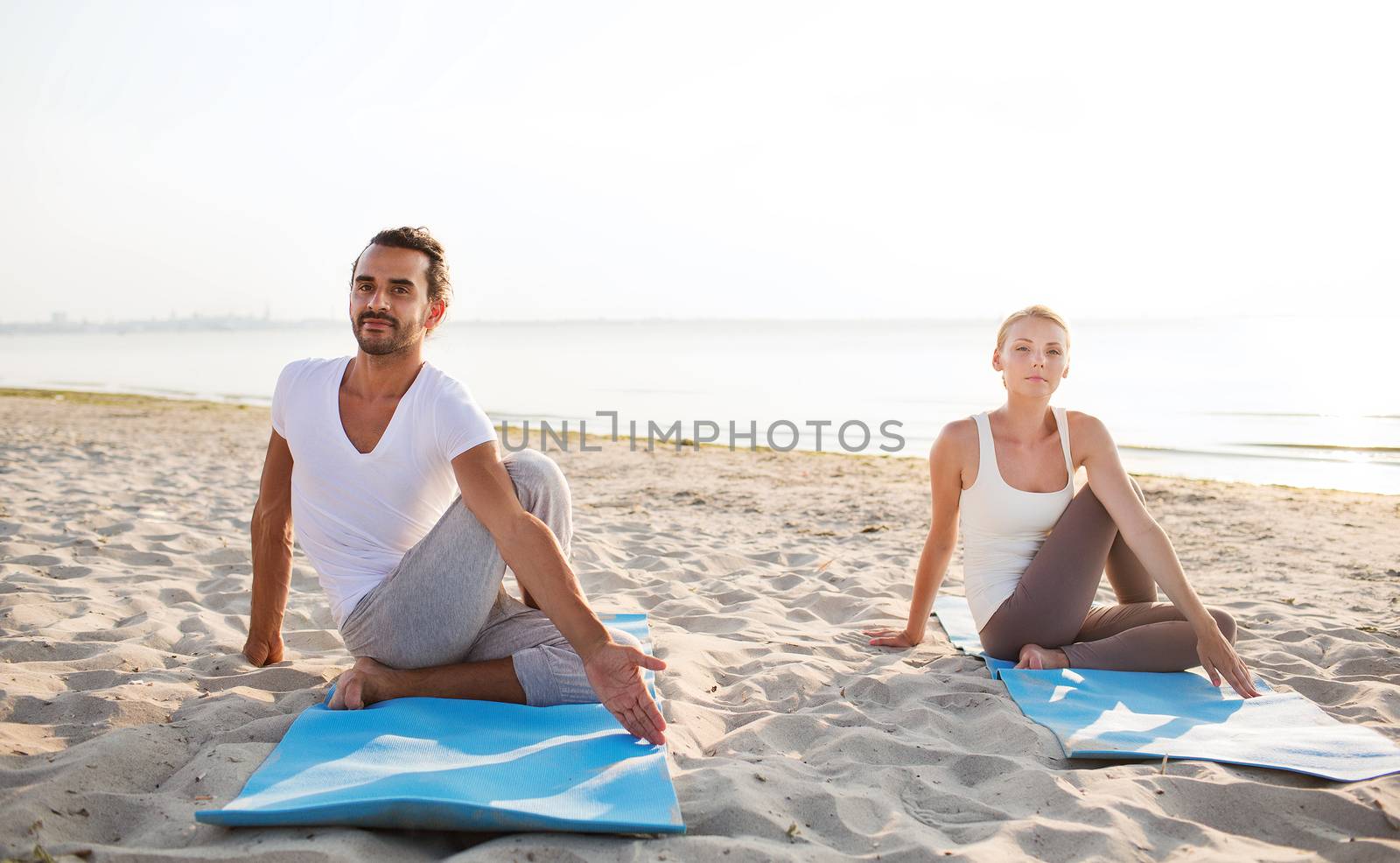fitness, sport, friendship and lifestyle concept - couple making yoga exercises sitting on mats outdoors