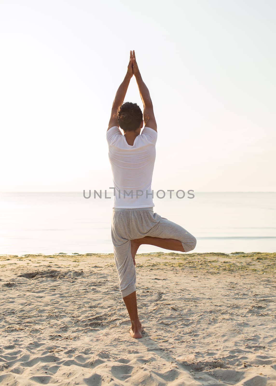 fitness, sport, people and lifestyle concept - man making yoga exercises on sand outdoors from back