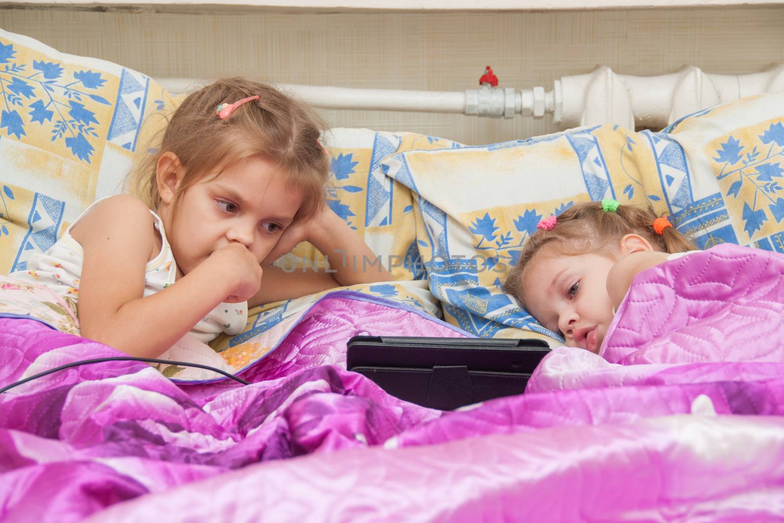 Two girls of three and five years, lie in bed and watch cartoons in the Tablet PC