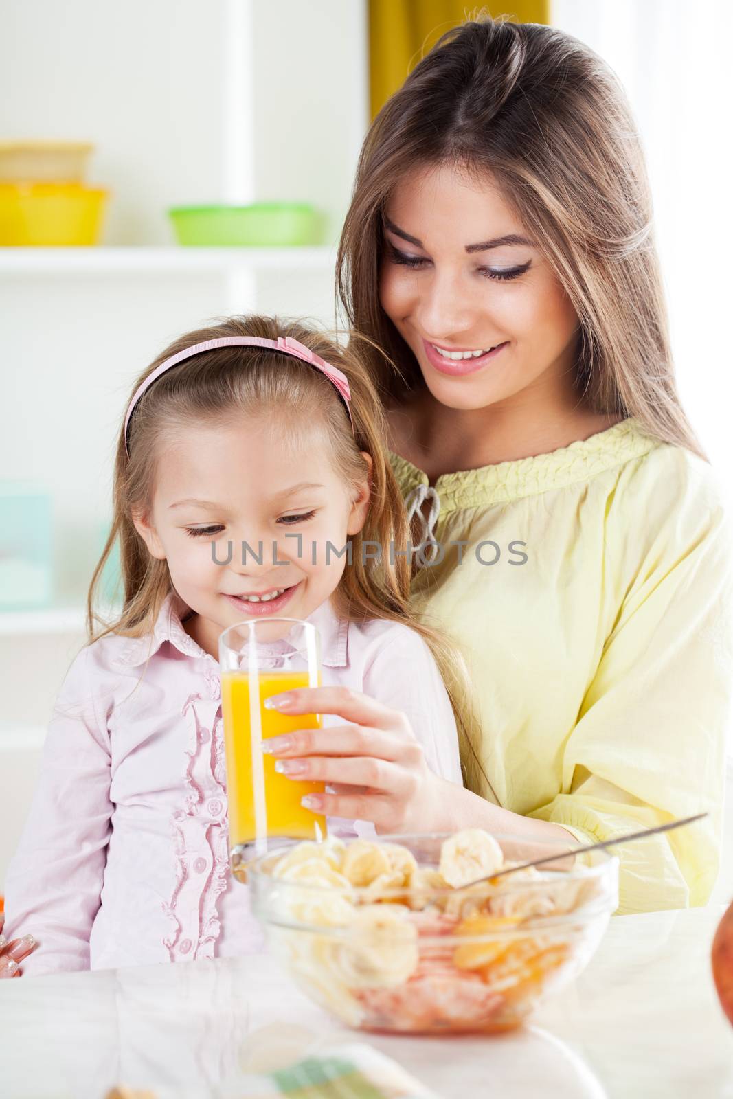 Beautiful young woman gives to the little girl orange juice and fruit salad in the kitchen.
