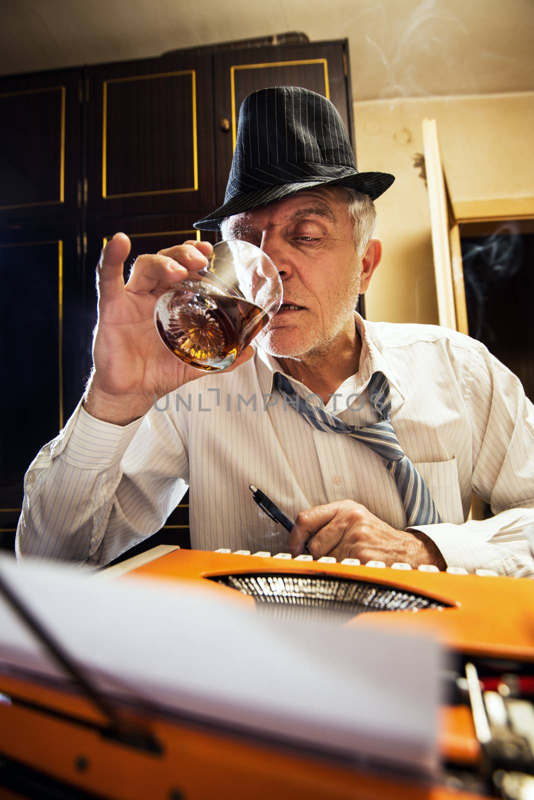 Retro Senior man, journalist, writer, writing on a traditional Typewriter. He is sitting with a glass of whiskey.