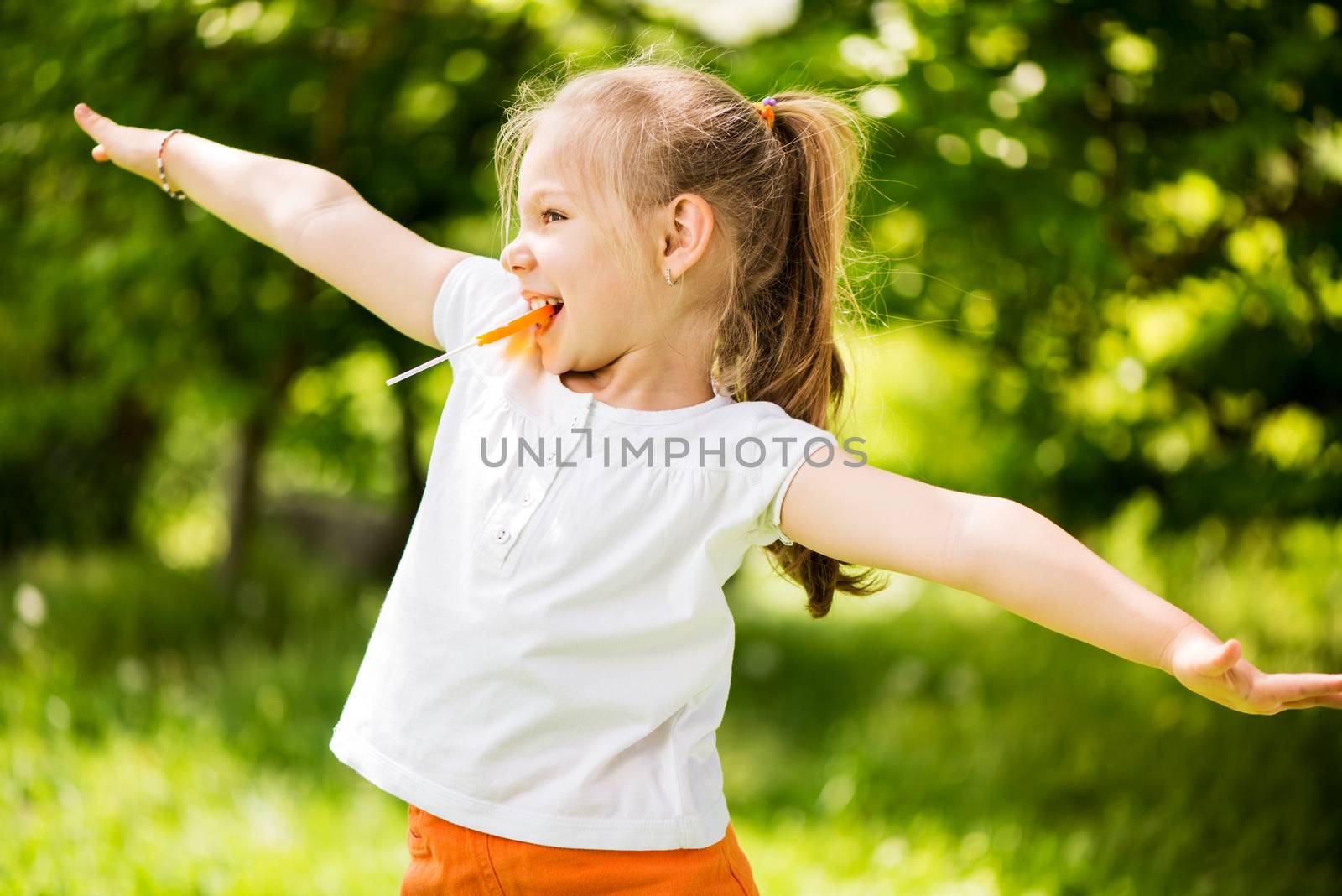 Cute Little Girl having fun in The Park and holding in mouth orange Lollipop.