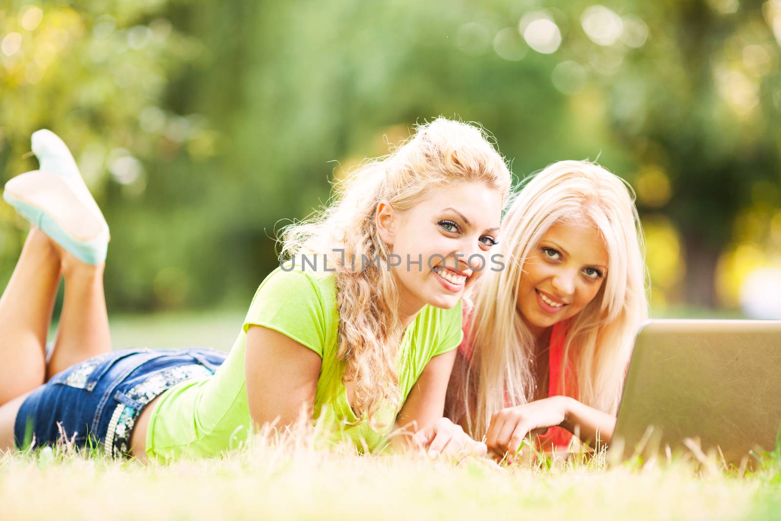 Two Beautiful girls lying in the grass with laptop and surfing on the internet.