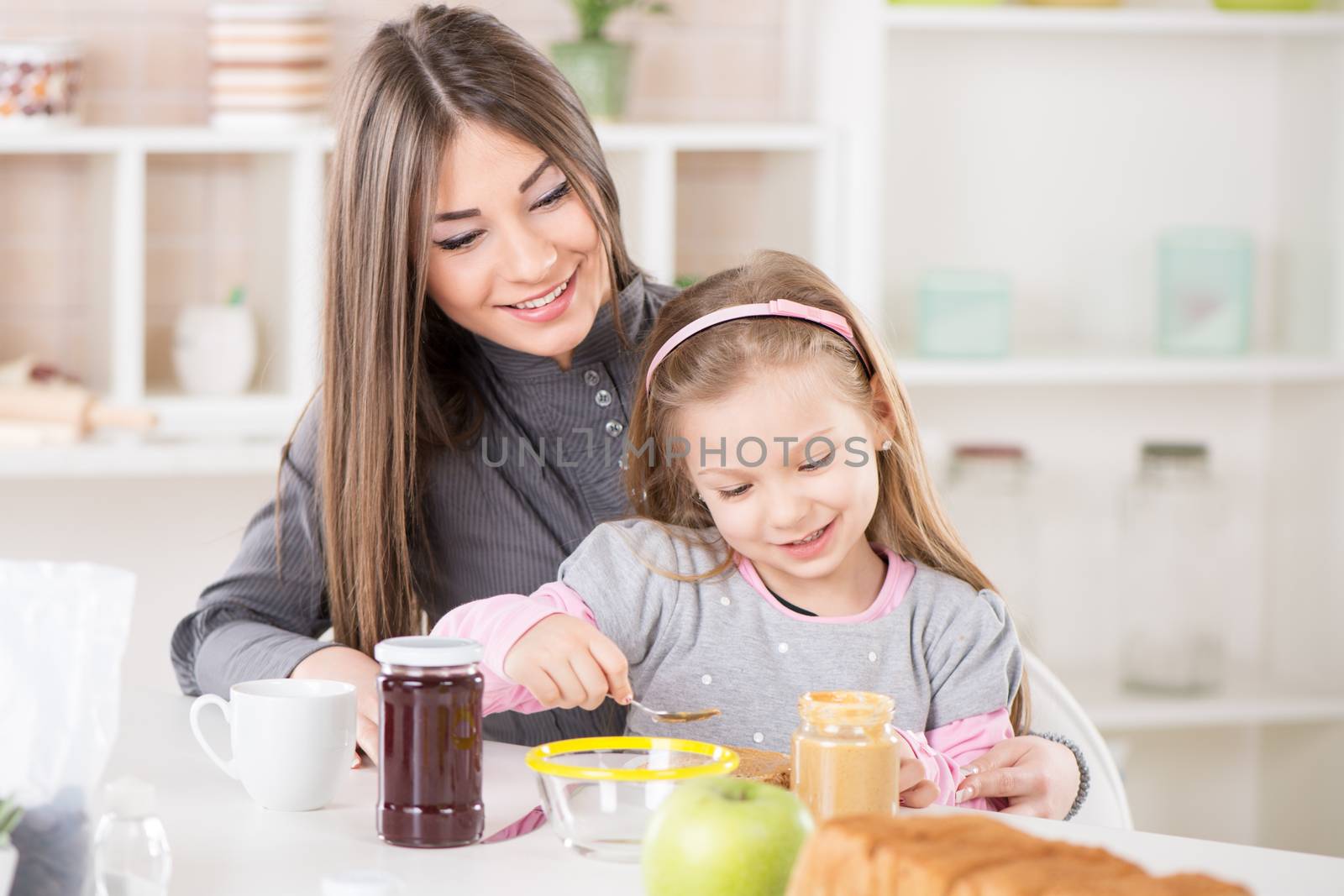 Cute little girl sitting in the mother lap and smearing peanut butter on bread.