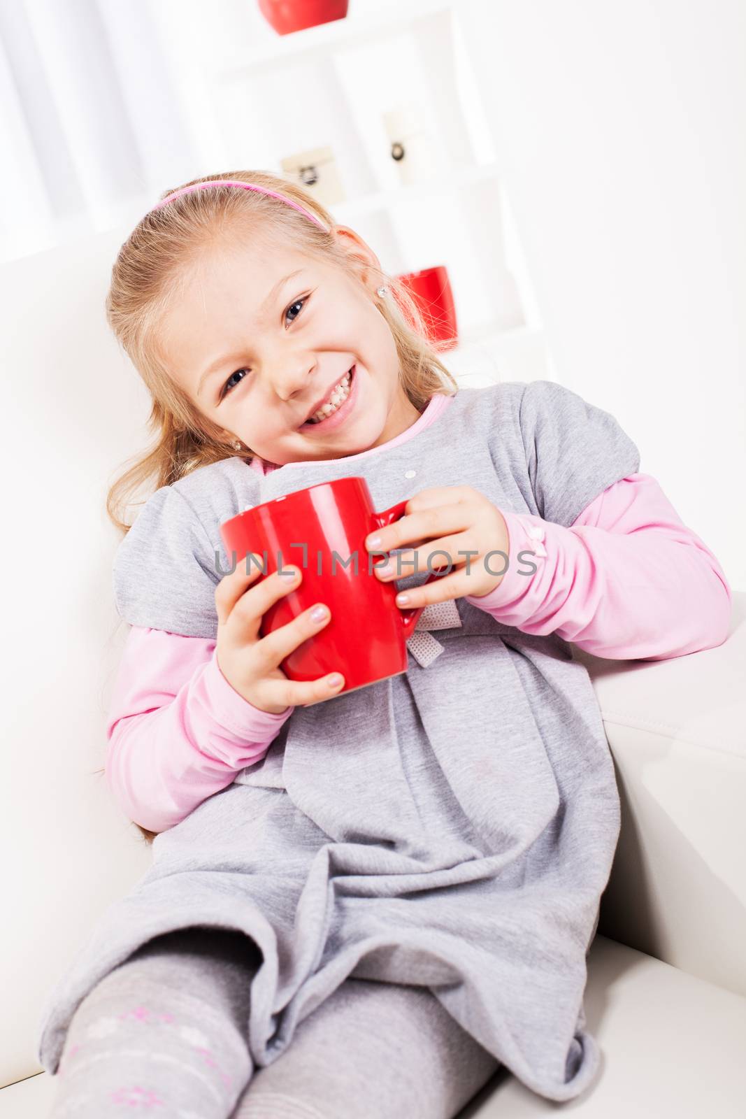 Cute little girl drinking tea from a red cup at home.