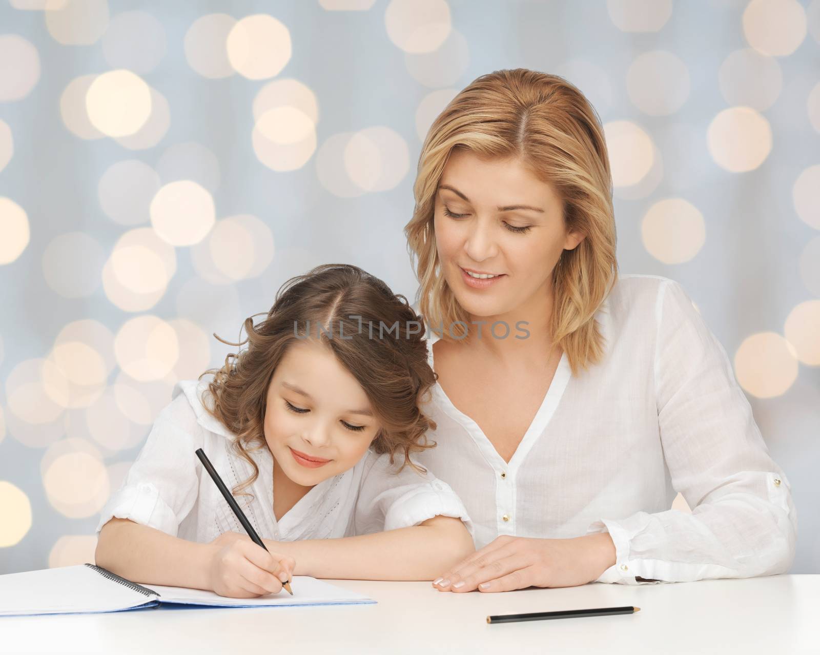 people, family, school, home education and parenting concept -happy mother and daughter doing homework and writing in notebook over holidays lights background