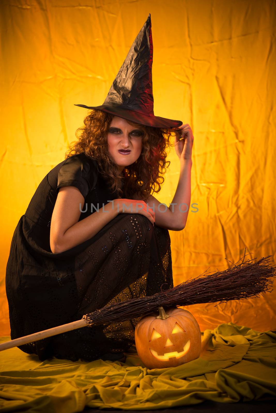 Young woman dressed like a witch. She is in dark clothing with broom and pumpkin.