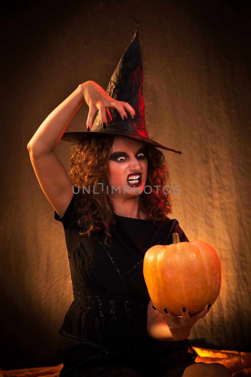 Young woman with evil face dressed like a witch. She wears dark clothing and holding a pumpkin in hand.