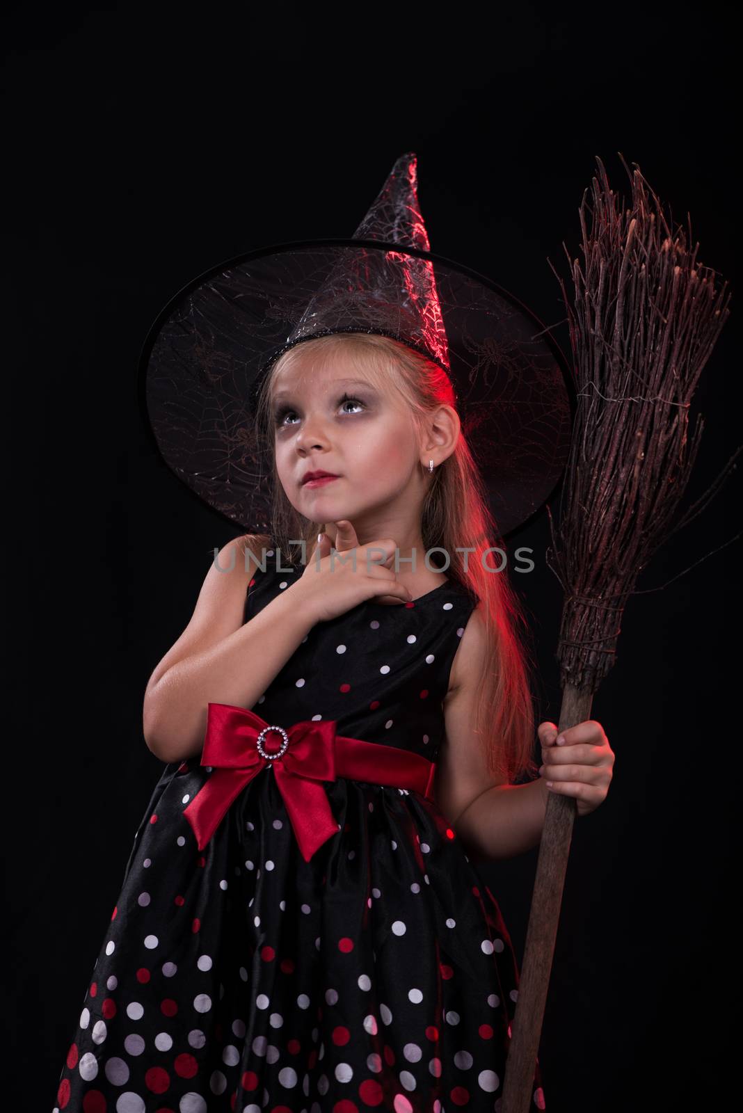 Little girl in dark clothing with broom on black background. She thinks.