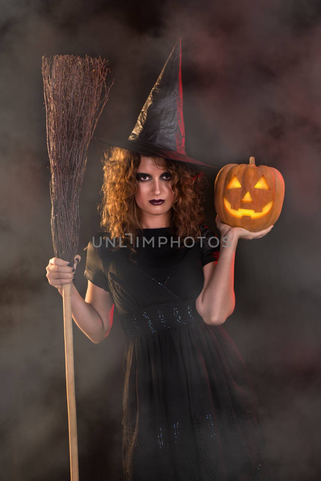 Young woman dressed like a witch. She is in dark clothing and holding broom and pumpkin.