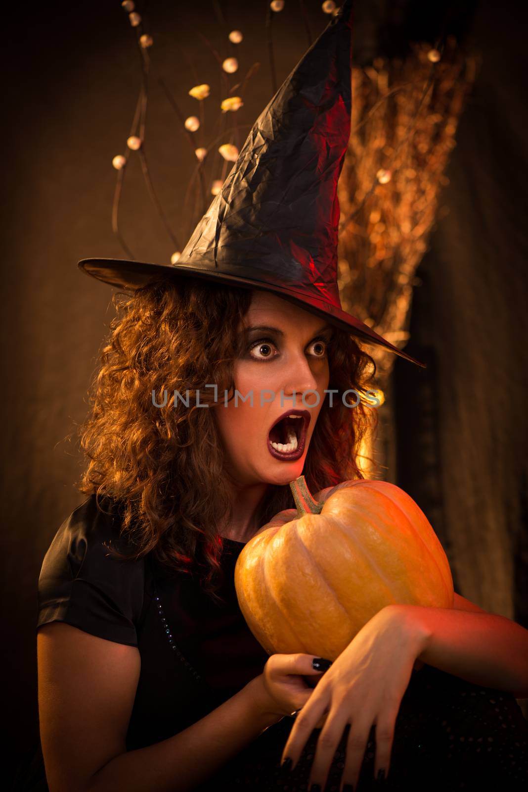 Portrait of young woman with scared face dressed like a witch. She wears dark clothing and holding a pumpkin in hands.